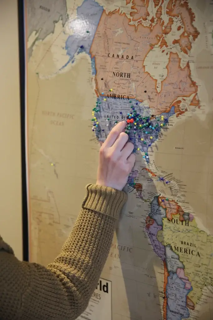 A hand places a push pin into the portion of a world map that includes North and South America. Several push pins are already placed in a variety of locations.